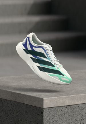Witte sneaker met zwarte, blauwe en groene accenten zwevend boven een betonnen richel tegen een wazige grijze achtergrond.