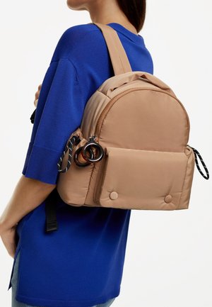 Mochila de nylon marrón con un diseño redondeado, bolsillo frontal, botones de presión y acentos de hardware negro, llevada sobre una camiseta azul.