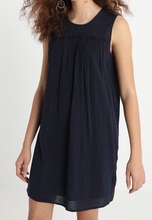 Donna che indossa un vestito senza maniche, ampio, di colore blu scuro navy con dettagli plissettati sotto la scollatura e orecchini a cerchio, mostrata dalle spalle fino a metà coscia.