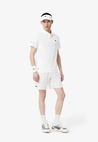 Vit sportpolo med korta ärmar och meshdetaljer, tillsammans med vita shorts. Har en grön logotyp. Bärs med en vit visir och atletiska skor.