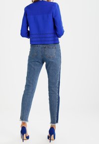 Kvinna klädd i en blå perforerad jacka, tvåtonade blå jeans med fransade fållar och matchande blå höga klackar, stående vänd bort.