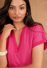 Top rosa brillante con escote en V y mangas enrolladas. Modelo lleva una pulsera gruesa de color claro. Textura suave, diseño minimalista.