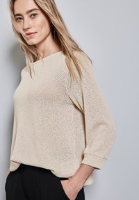 Maglione lavorato a maglia di un beige chiaro, con una vestibilità ampia, scollatura larga e maniche a tre quarti. La texture è leggera e ariosa, offrendo un aspetto rilassato.