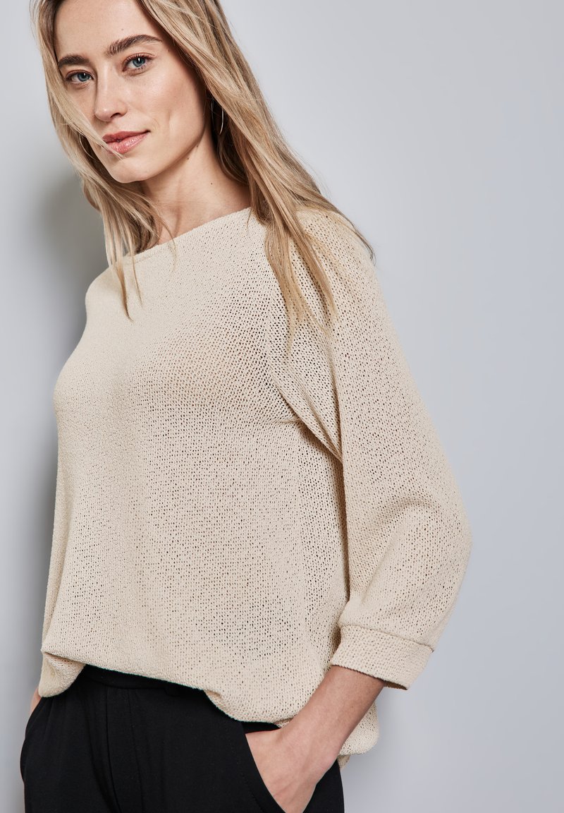 Maglione lavorato a maglia di un beige chiaro, con una vestibilità ampia, scollatura larga e maniche a tre quarti. La texture è leggera e ariosa, offrendo un aspetto rilassato.