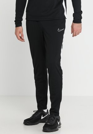 Mand iført sorte Nike-træningsbukser og sorte Nike-sneakers med hvide swoosh-logoer, stående mod en ensfarvet baggrund.