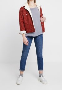 Chaqueta de mezclilla roja con cuello de piel sintética blanca, camiseta gris, jeans azules y zapatillas blancas. La chaqueta cuenta con bolsillos y mangas enrolladas.