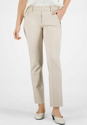 Beige op maat gemaakte broek met zijritszakken, gedragen met witte muiltjes met hak, model van taille tot voeten gecropped.