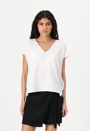 Blouse blanche à manches courtes et décolleté en V en tissu léger ; présente une coupe décontractée avec un ourlet légèrement arrondi ; portée sur un short noir.