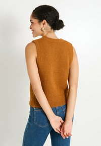 Femme aux cheveux bouclés en chignon portant des boucles d'oreilles en or, un haut en tricot sans manches marron et un jean bleu, se tenant avec les mains derrière le dos.