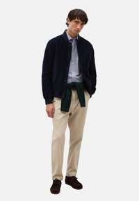 Giubbotto bomber in corduroy blu navy, camicia a righe azzurro chiaro, pantaloni beige, maglione verde annodato in vita, scarpe in suede marrone, design minimalista.