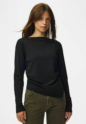 Femme aux longs cheveux bruns portant un haut noir à manches longues et un pantalon vert olive, debout devant un fond blanc uni.