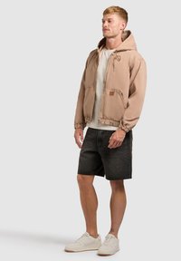Homme portant une veste à capuche beige, un t-shirt blanc, un short en jean noir et des baskets blanches, debout avec une expression neutre sur un fond gris.