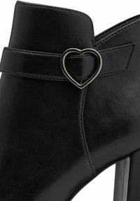 Schwarze Leder-Ankle-Boots mit einem herzförmigen silbernen Akzent an einem Riemen, schlanker Form, glatter Textur und hohen Absatz.