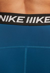 Blå idrottsshorts med en svart midja som har Nike-logotypen i vitt. Slät textur med sömmar synliga längs botten.