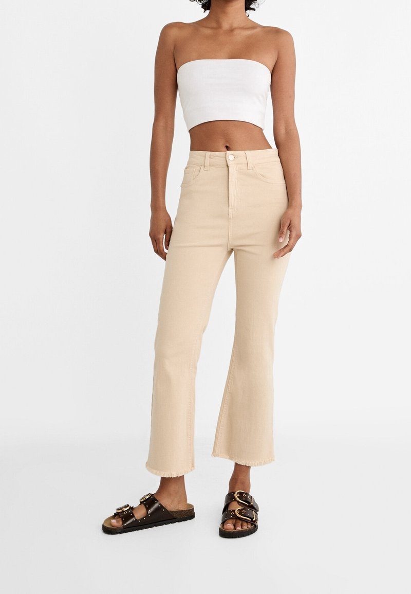 Jean flare beige avec ourlets effilochés, taille haute et fermeture à boutons devant, assorti à un haut blanc sans bretelles et des sandales marron à enfiler.