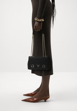 Personne portant une robe midi marron côtelée tenant un sac à main noir avec des chaînes dorées, portant des chaussures à talons kitten marron à bout pointu et un bracelet épais en or.