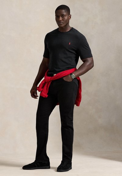 Černé tričko s krátkým rukávem s červeným logem, doplněné o černé džíny a černé tenisky. Červený svetr uvázaný kolem pasu přidává barevný kontrast.