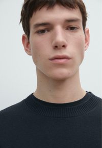 Maglione scuro a trama con scollo rotondo a coste. I lineamenti del volto del modello sono visibili su uno sfondo chiaro.