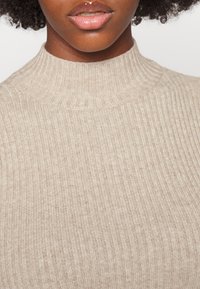 Beige tröja med hög ribbad krage, som har ett strukturerat stickat mönster. Materialet verkar mjukt och något elastiskt.