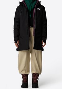 Chaqueta aislante negra con capucha, forro morado, llevada sobre un suéter verde. Combinada con pantalones beige y botas de cordones rojo oscuro.