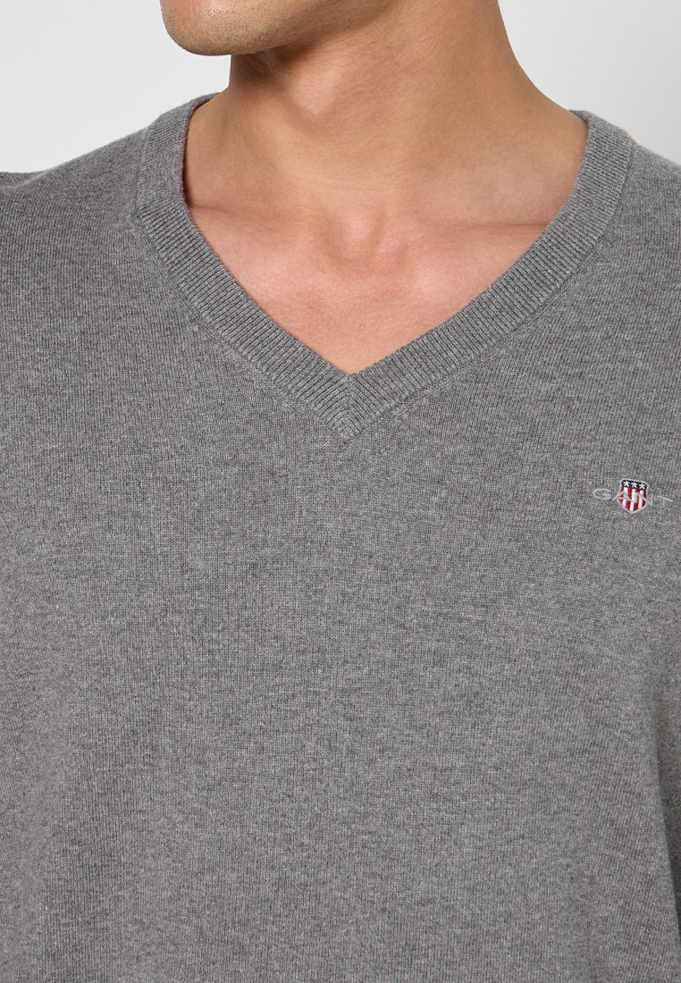Grauer V-Ausschnitt-Pullover mit einer weichen Textur. Verfügt über einen gerippten Ausschnitt und ein kleines Logo-Emblem auf der linken Brust.