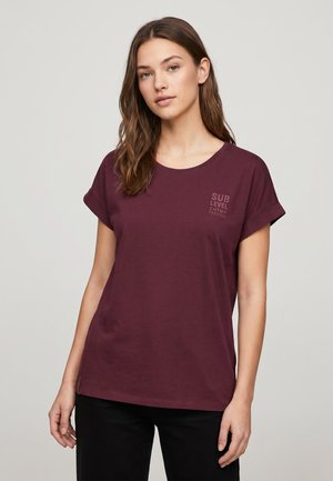 Junge Frau mit langen braunen Haaren in einem dunkelroten kurzärmeligen T-Shirt mit kleinem Text auf der linken Brust und schwarzen Hosen vor einem schlichten Hintergrund.