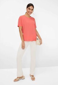 Coral short-sleeve top with a round neckline, paired with white flared pants and beige sandals. Model holding a small cream handbag.