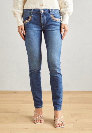Mujer con vaqueros pitillo azules con detalles de bolsillos de estampado de leopardo, suéter crema y sandalias de tacón blancas con tiras, de pie sobre suelo de madera.