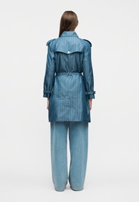 Γυναίκα, φαίνεται από πίσω, φοράει καμπαρντίνα με μπλε μοτίβα και ζώνη, με επιμανίκια και φαρδιά ανοιχτό μπλε τζιν παντελόνια, σε απλό λευκό φόντο.