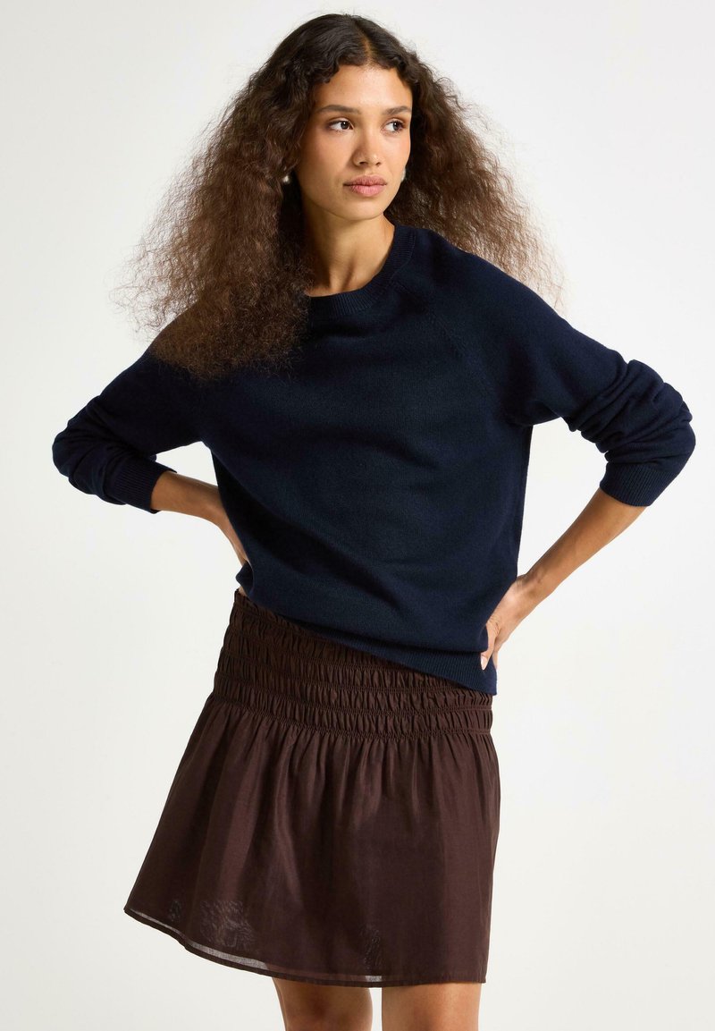 Maglione lavorato a maglia blu navy con scollo rotondo e maniche lunghe, abbinato a una gonna corta marrone con vita smock.