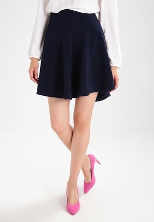 Vrouw draagt een marineblauwe uitlopende rok, witte blouse met lange mouwen en roze hoge hakken, staand met gekruiste benen op een witte achtergrond.