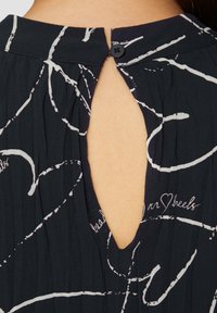 Tissu noir avec des motifs abstraits blancs, comportant une ouverture en forme de clé dans le dos, sécurisée par un petit bouton noir. Texture plissée sur l'ensemble.