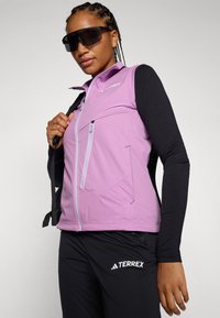 Jachetă mov fără mâneci, cu fermoar în față, două buzunare laterale și accente texturate, purtată peste un top negru cu mânecă lungă. Pantaloni negri cu logo.