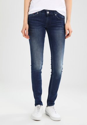 Personne portant un jean slim bleu foncé et des baskets blanches, debout devant un fond clair uni.