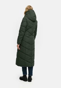 Lång, grön puffjacka med huva, som har ett diamant-quiltat mönster. Den har långa ärmar och når under knäna. Stylad med svarta stövlar.