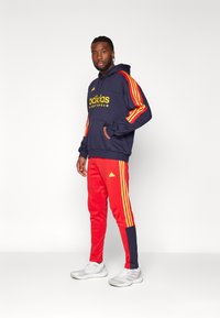 Felpa della marina con logo "adidas" giallo e strisce rosse sulle maniche; abbinata a pantaloni rossi con striscia laterale e logo; scarpe sportive grigie.