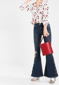 Blouse blanche avec des motifs floraux rouges et des pois noirs, associée à un jean évasé foncé avec une déchirure et des boutons. Tenant un sac seau rouge.