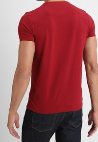 Mand iført ensfarvet rød t-shirt med korte ærmer og mørkeblå jeans, vist fra ryggen mod en hvid baggrund.