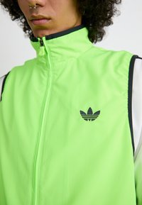 Neon zöld sportmellény magas gallérral, cipzáros záródással és fekete adidas háromlomb logóval. Fekete díszítéssel rendelkezik a karmegnyitóknál.