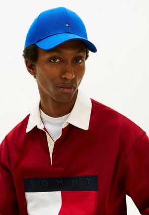 Jeune homme portant une casquette bleue et un polo rouge avec un col blanc et un grand logo Tommy sur la poitrine, sur un fond uni.