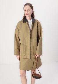 Kaki jacka med mörk krage, har stora framfickor och dragkedjestängning. Modellen håller en liten brun handväska.