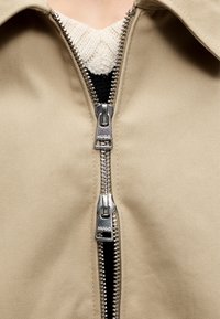 Gros plan d'une veste beige avec deux fermetures éclair argentées HUGO partiellement ouvertes, révélant un tricot noir et blanc en dessous.