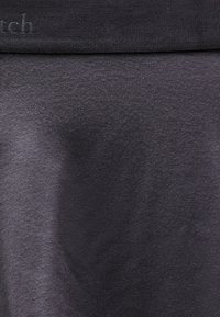 Κοντινό πλάνο σκουρόχρωμου γκρι υφάσματος με διακριτική υφή, που δείχνει αμυδρές σκιές και μερικό, θολό ανάγλυφο κείμενο στην επάνω αριστερή γωνία.