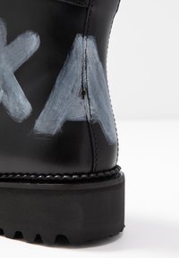 Zwarte leren laars met zichtbare grijze verfletters. Voorzien van een dikke, gestructureerde zool en stiksels rond de basis. Over het geheel genomen een glad oppervlak.