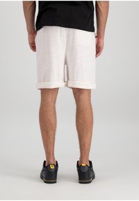 Shorts blancs texturés avec des revers et une poche arrière, associés à des chaussures noires avec des accents jaunes. Vue de derrière.