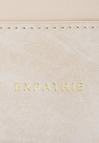 Világos bézs textúrájú bőr, középen, az alján arany felirattal "EXPATRIÉ" betűzve. Finom varrás a széleken.