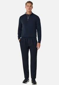Pull en tricot bleu marine avec col montant et fermeture éclair, associé à un pantalon sombre. Le matériau semble lisse et doux avec un design à coupe décontractée.