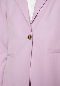 Světle fialový blazer vyrobený z hladkého materiálu, s jedním kulatým zlatým knoflíkem a dvěma vertikálními kapsami. Střih je na míru s ostrými klopami.