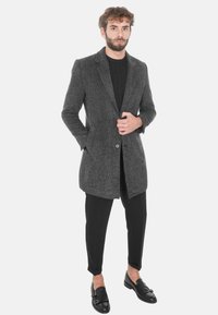 Cappotto lungo grigio da uomo in tessuto testurizzato, con rever a risvolto, chiusura con un solo bottone e tasche laterali, abbinato a pantaloni neri e scarpe.