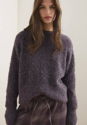 Pull en mohair violet, avec une coupe décontractée, épaules tombantes et une ourlet légèrement courbé sur les côtés. Porté par-dessus une jupe à carreaux.